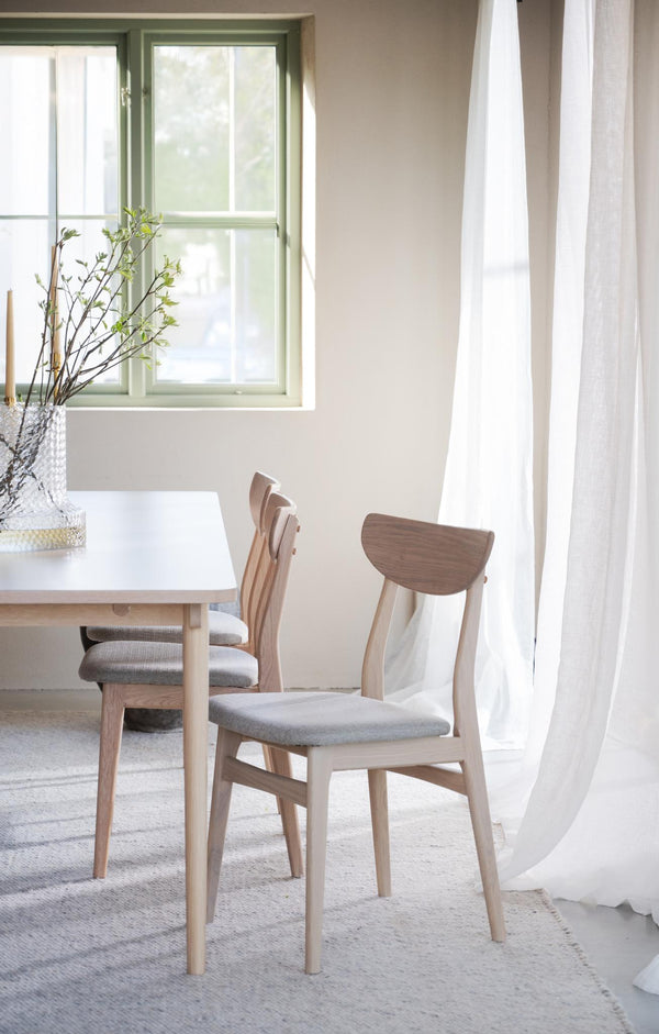 Matgrupp med stolar i ljust trä och grå dynor vid fönster med vita gardiner i ljust matrum