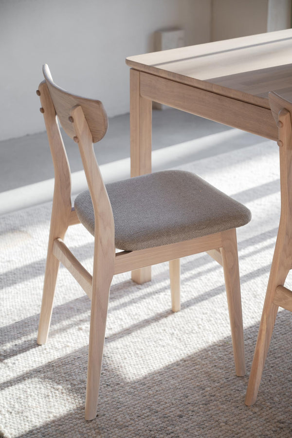 Matstol i ljust trä med grå klädsel och rundad rygg vid matbord i skandinavisk design