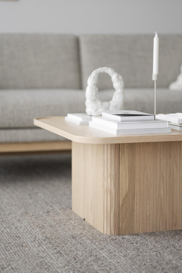 Ljust träsoffbord med räfflad kant, dekorerat med vita böcker, vit skulptur och ljusstake framför grå soffa