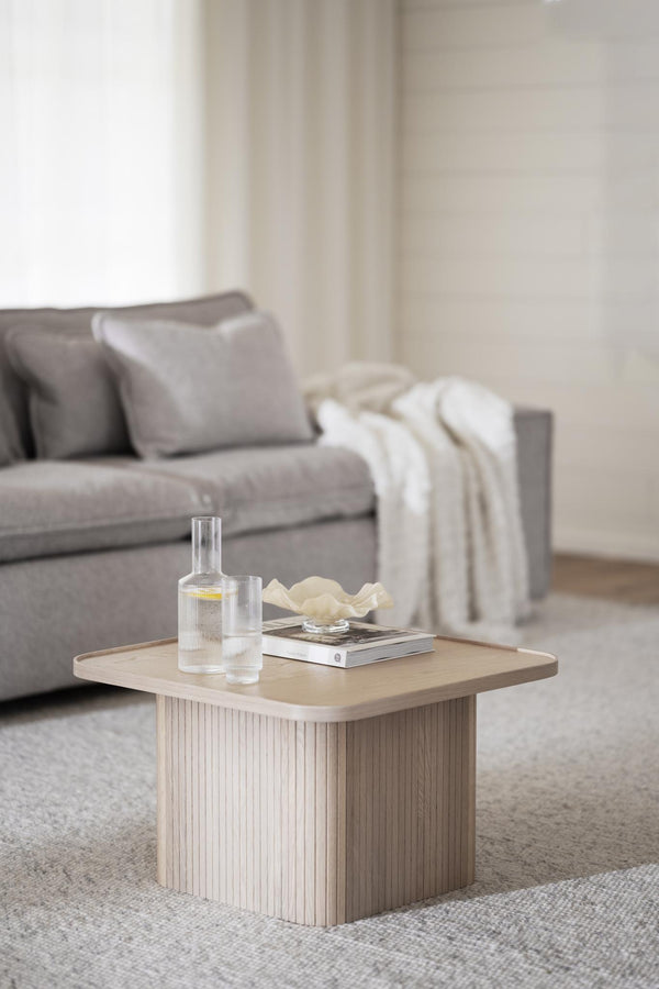 Ljust soffbord i trä med räfflad bas, dekorerat med glaskaraff, böcker och vit skål i modern vardagsrumsmiljö