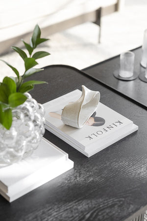 Vit keramisk dekorationsvas i form av blad på coffeetable-bok bredvid grön växt och glasvas