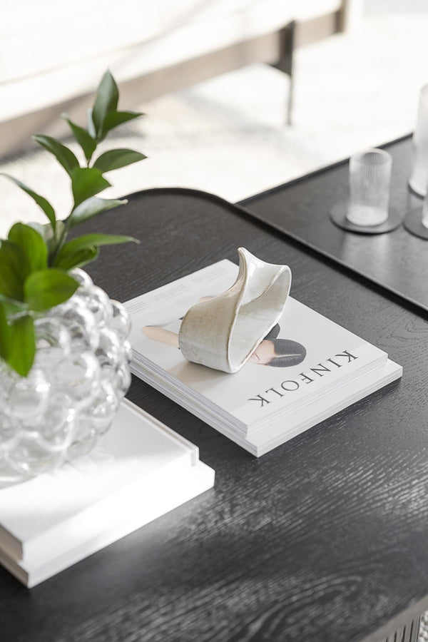 Vit keramisk dekorationsvas i form av blad på coffeetable-bok bredvid grön växt och glasvas