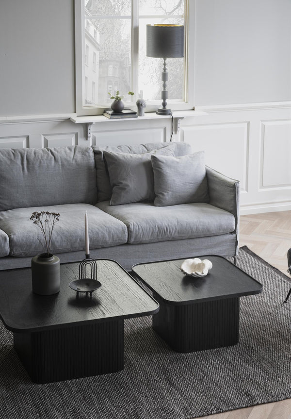 Grå linsoffa med kuddar framför soffbord i svart trä i skandinaviskt vardagsrum med vit väggpanel