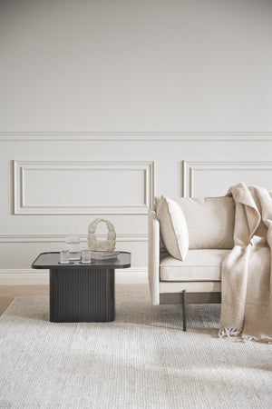 Beige fåtölj med vit kudde och pläd bredvid svart soffbord med dekor i elegant vardagsrum