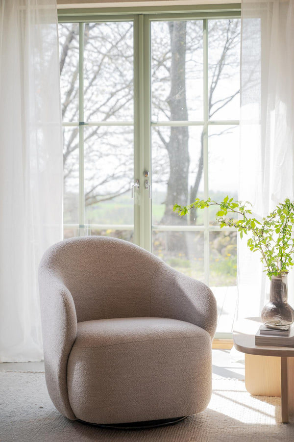Beige bouclé fåtölj vid fönster med transparenta gardiner och gröna karmar, bredvid runt träbord med glasvas