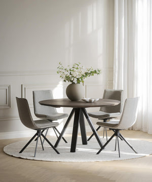 Runt matbord i mörkt trä med fyra gråa stolar och svarta metallben i ljust matrum med vita väggar