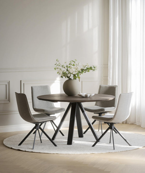 Runt matbord i mörkt trä med fyra gråa stolar och svarta metallben i ljust matrum med vita väggar
