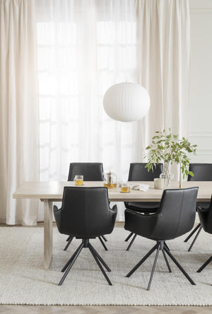 Matbord i ljust trä med svarta stolar i skinn, vit taklampa och genomskinliga gardiner i modern matsal