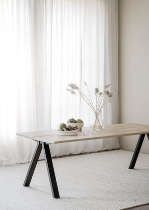 Matbord med ljust trä och svarta A-ben, dekorerat med vaser och växter framför vita gardiner