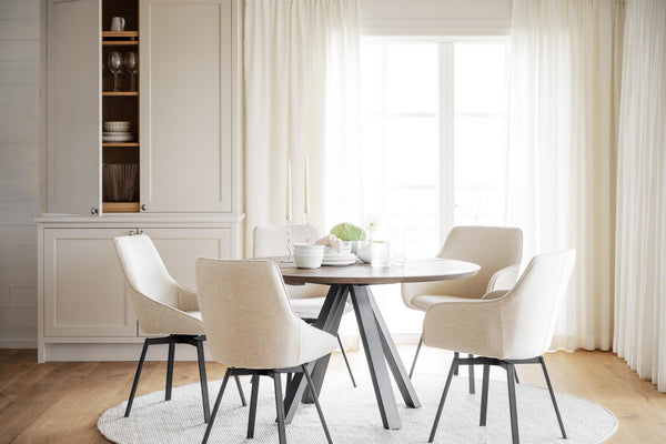 Matgrupp med runt bord och beige stolar i ljus matsal med vitrinskåp och gardin