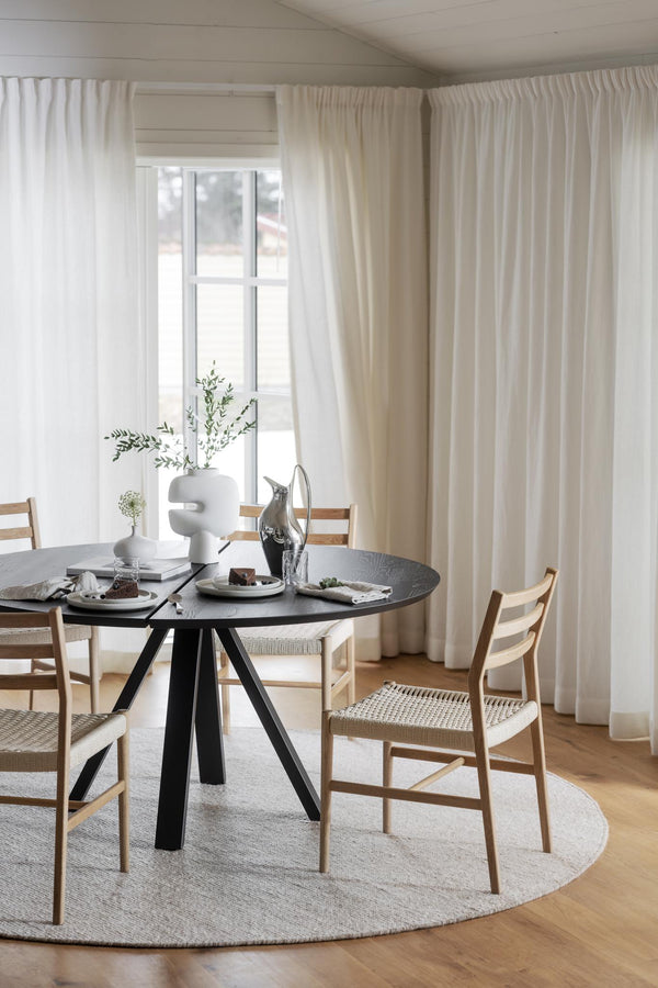 Rund svart matbord med ljusa trästolar i skandinavisk matsal med beiga gardiner och fönster