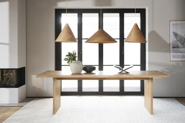 Matbord i ljust trä med tre rotting taklampar ovanför i modern matsal med svarta fönsterpartier