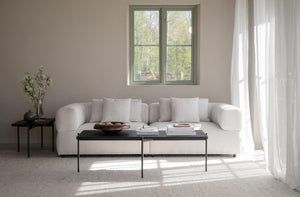 Minimalistiskt vardagsrum med beige soffa, svart soffbord och ljusa textilier i skandinavisk stil