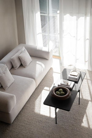 Minimalistiskt vardagsrum med beige soffa, vit gardin och svart soffbord med dekorativa böcker och skål