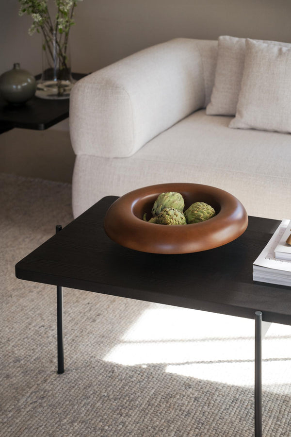 Terrakottafärgad dekorativ skål med gröna artiskockor på svart soffbord framför beige soffa