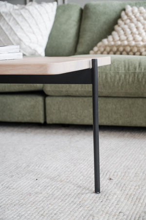 Soffbord med ljus träskiva och svarta metallben framför grön sammetssoffa med kuddar