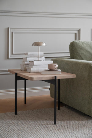 Soffbord i ljust trä med svarta ben bredvid grön fåtölj, dekorerat med böcker och bordslampa