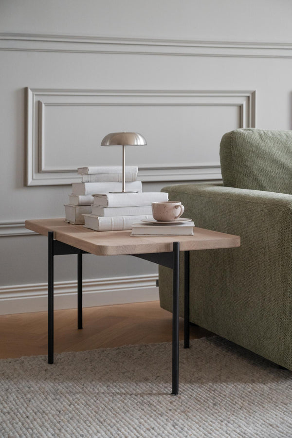 Soffbord i ljust trä med svarta ben bredvid grön fåtölj, dekorerat med böcker och bordslampa