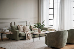 Grönt sammetstyg soffa med beige kuddar och pläd i ljust vardagsrum med fönster och vita gardiner