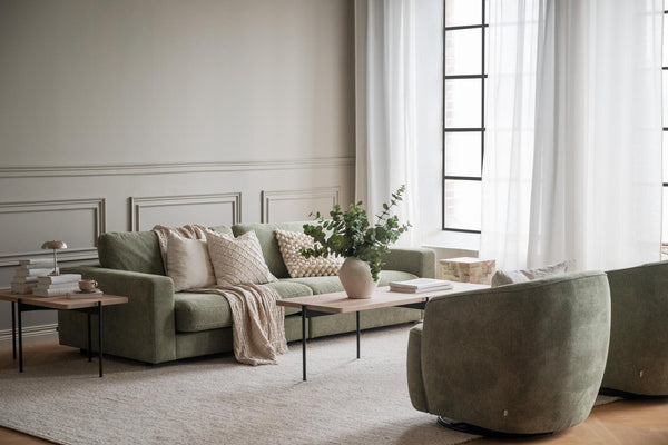 Grönt sammetstyg soffa med beige kuddar och pläd i ljust vardagsrum med fönster och vita gardiner