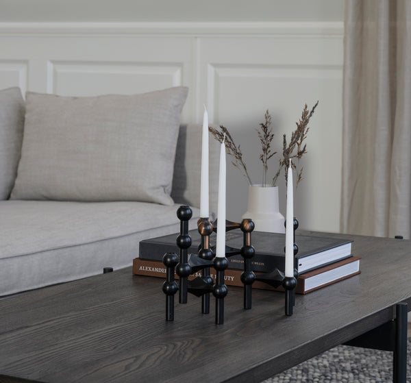 Svart ljusstake med molekyldesign och två vita ljus på träbord i vardagsrum med böcker och vas