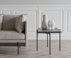 Grå sidobord med svarta ben bredvid beige sammetssoffa med vita keramiska vaser som dekoration