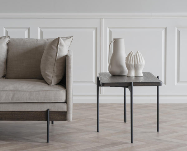 Grå sidobord med svarta ben bredvid beige sammetssoffa med vita keramiska vaser som dekoration