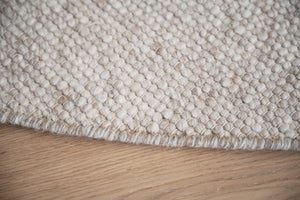 Närbild av ljus handvävd matta med strukturerad yta i beige och vita toner på trägolv