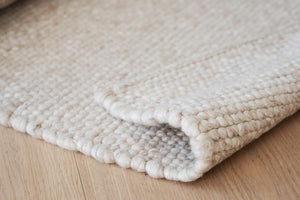 Närbild på hörnet av en handvävd matta i naturliga beige och vita toner på trägolv