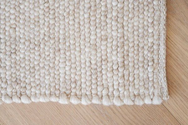 Handvävd matta i ljusbeige ull med strukturerad yta och tofsar, lagd på trägolv