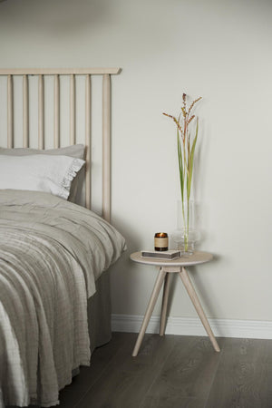 Ljust sovrum med beige sänggavel, grå linnelakan och runt sängbord i trä med vas och dekoration