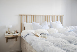 Säng med sängkläder i ljusblått och vitt, träsängavel med spjälor, sänghylla med bok och kaffekopp