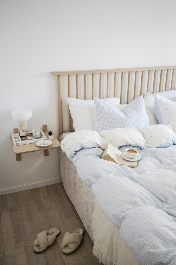 Ljust sovrum med ljust trä sängavel, blåvit rutigt sängkläder, kaffekopp på boken och tofflor på golvet