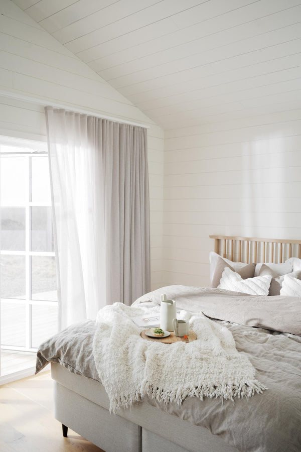 Ljust sovrum med vita sängkläder, beige pläd och gardiner i skandinavisk stil
