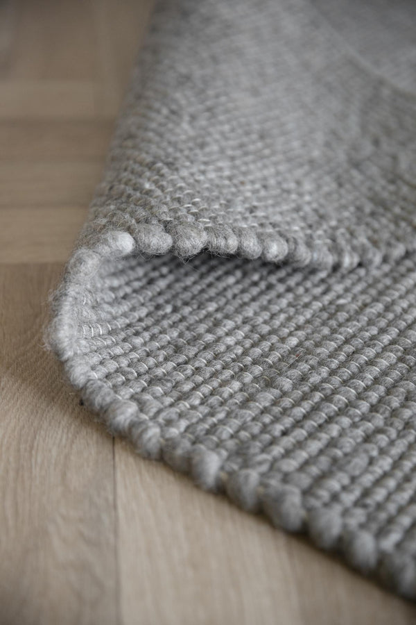 Grå handvävd ullmatta med strukturerad yta och synlig ovanlockskant, uppvikt på ljust trägolv