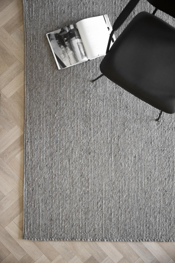 Grå ribbstickad matta på trägolv med svart stol och öppen bok