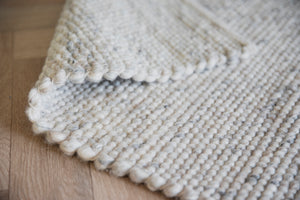 Närbild av stickad filt i ull med grå och vita toner, visar struktur och detaljer på trägolv