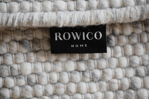 Närbild på ROWICO HOME-etikett i svart tyg mot beige stickad textil med fluffig vit kant