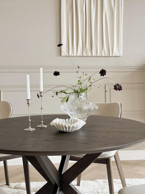 Runt matbord i mörkt trä med korsben och elegant blomsterarrangemang i kristallvas med ljusstakar