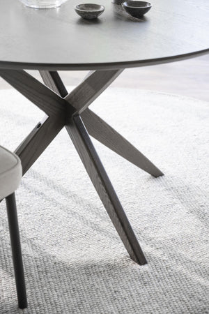 Runt matbord med grå träben i X-form och ljusgrå bordsskiva på vävd matta