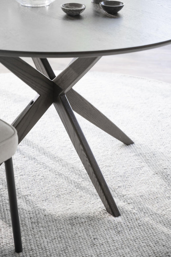 Runt matbord med grå träben i X-form och ljusgrå bordsskiva på vävd matta