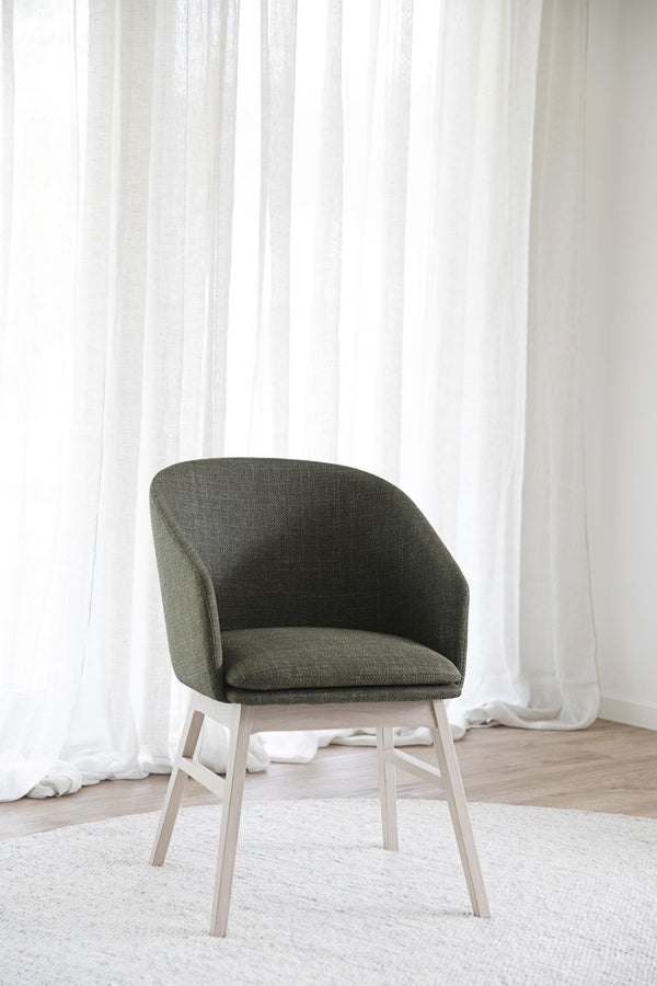 Grön stoppad stol med ljusa träben framför vita gardiner i minimalistisk skandinavisk stil