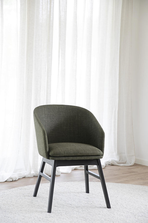 Grön stoppad stol med svarta ben framför vit gardin på ljust trägolv