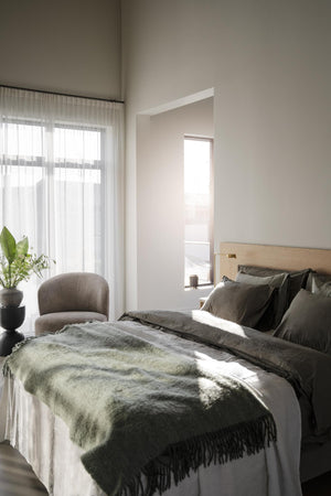 Ljust sovrum med olivgrön pläd och kuddar på säng, stor fönsterfönster med gardiner och gröna växter