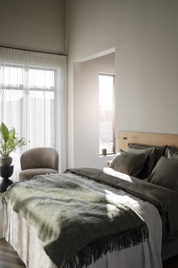 Ljust sovrum med olivgrön pläd och kuddar på säng, stor fönsterfönster med gardiner och gröna växter