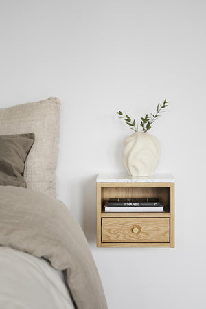 Sängbord i ek med låda och öppen hylla, vit marmor-topp, dekorerat med vas och böcker vid säng