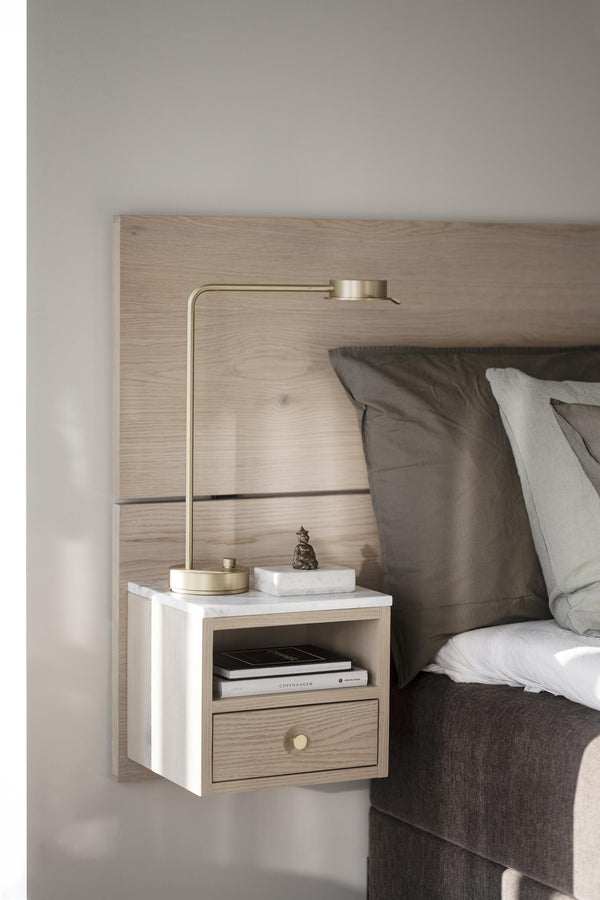 Väggmonterat nattduksbord i vit marmor och ljust trä med mässinglampa vid säng med grå sängkläder