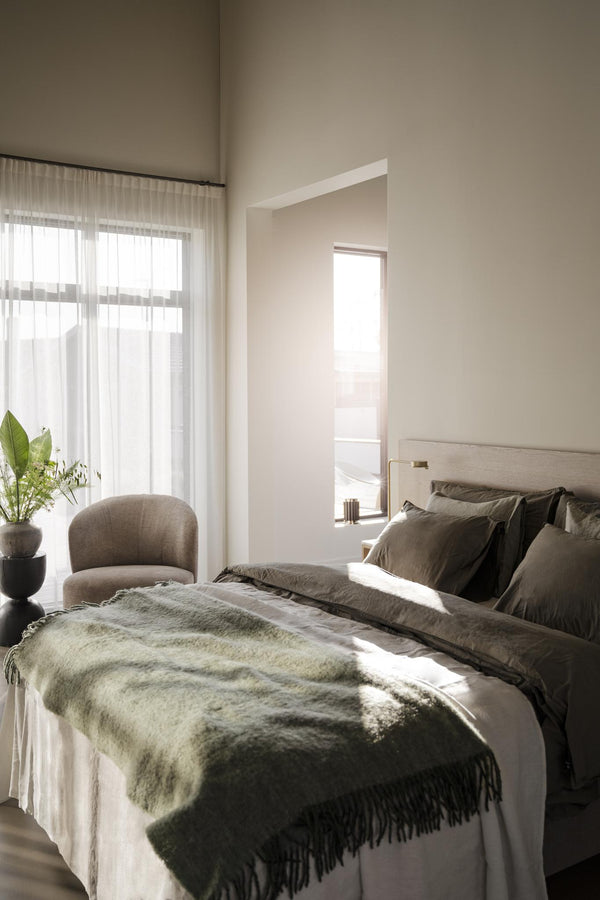 Ljust sovrum med beige sängkläder, grön pläd med fransar och vita genomskinliga gardiner vid fönster