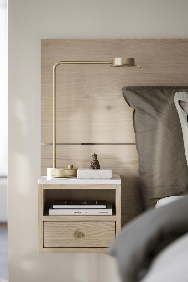 Sängbord i ljust trä med låda, guldlampa och gråa sängkuddar i sovrum med skandinavisk design