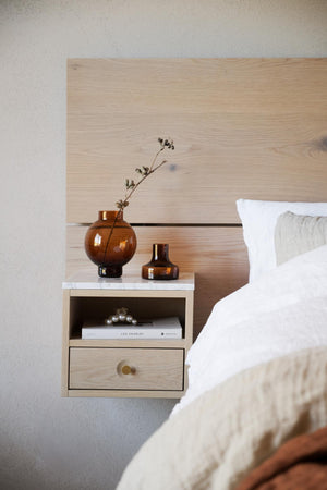 Vägghängt sängbord i ljust trä med låda och öppen hylla, dekorerat med amberbruna glasvaser och böcker